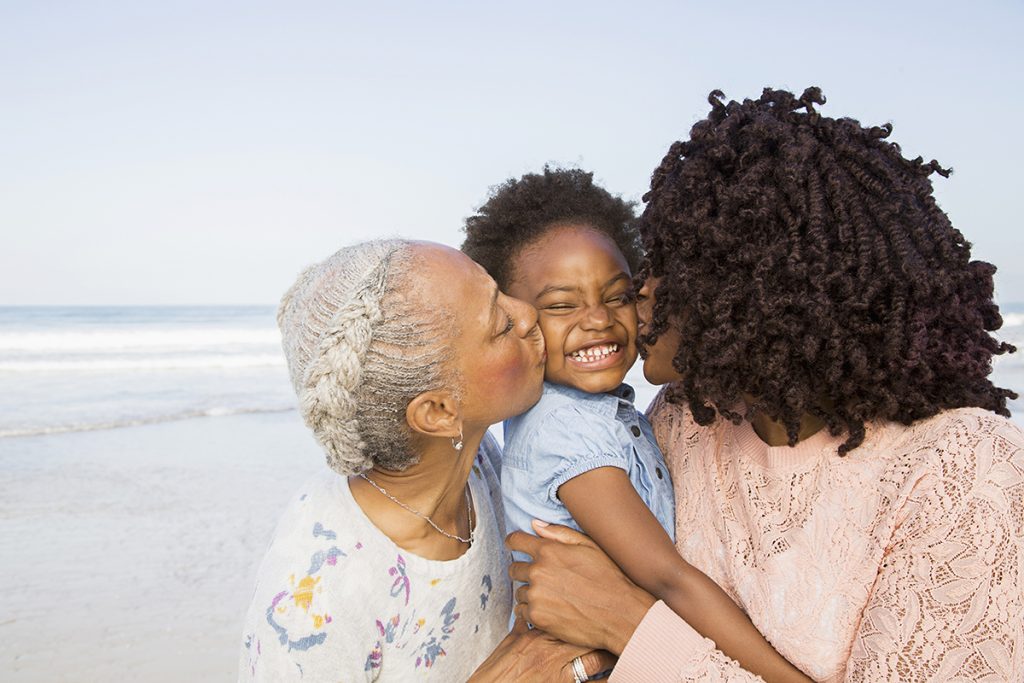 Black mother and grandmother kissing cheeks of girl on beach